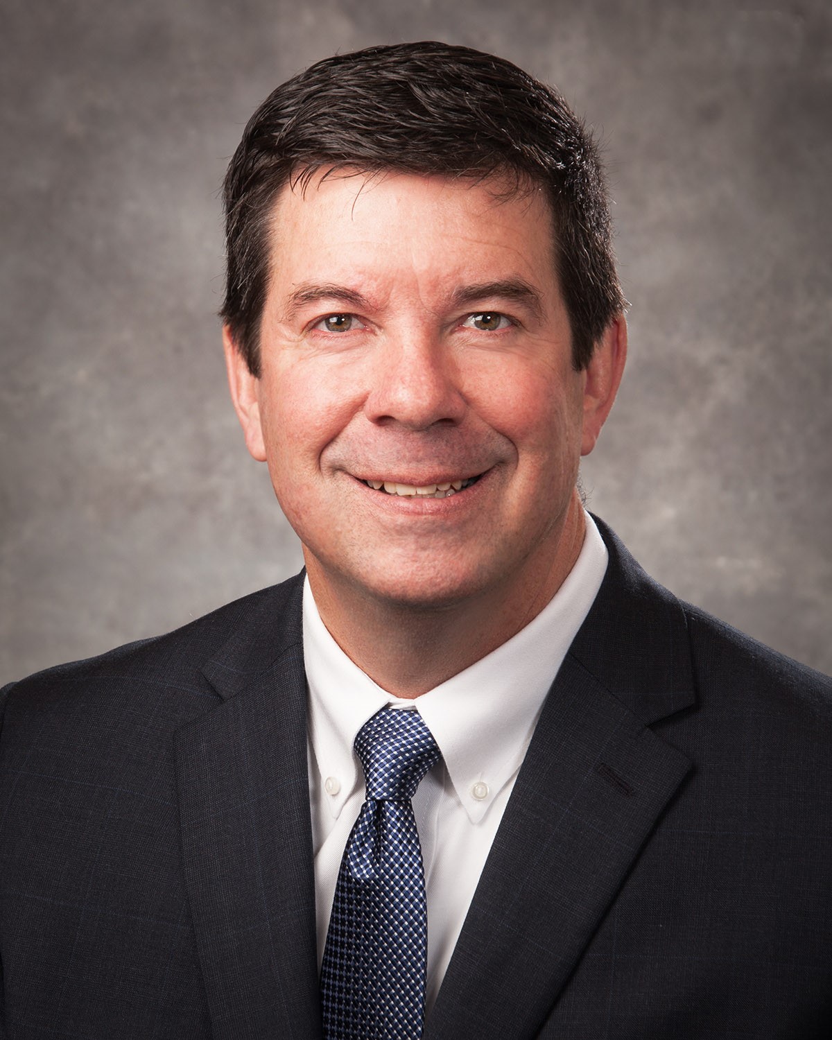 Smiling man in a dark suit, white shirt, and patterned blue tie