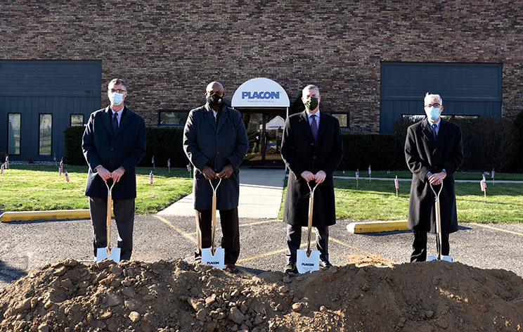 Four masked men in coats hold shovels at a groundbreaking ceremony outside a PLACON building