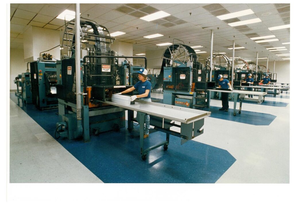 Workers operate industrial printing or packaging machines in a factory