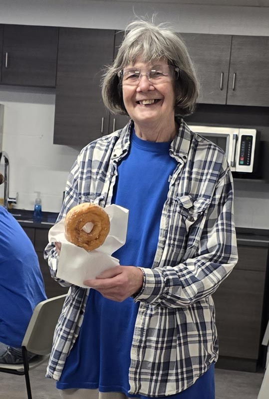 Careers 1 Smiling woman wearing glasses and a plaid shirt holds a bagel wrapped in a napkin