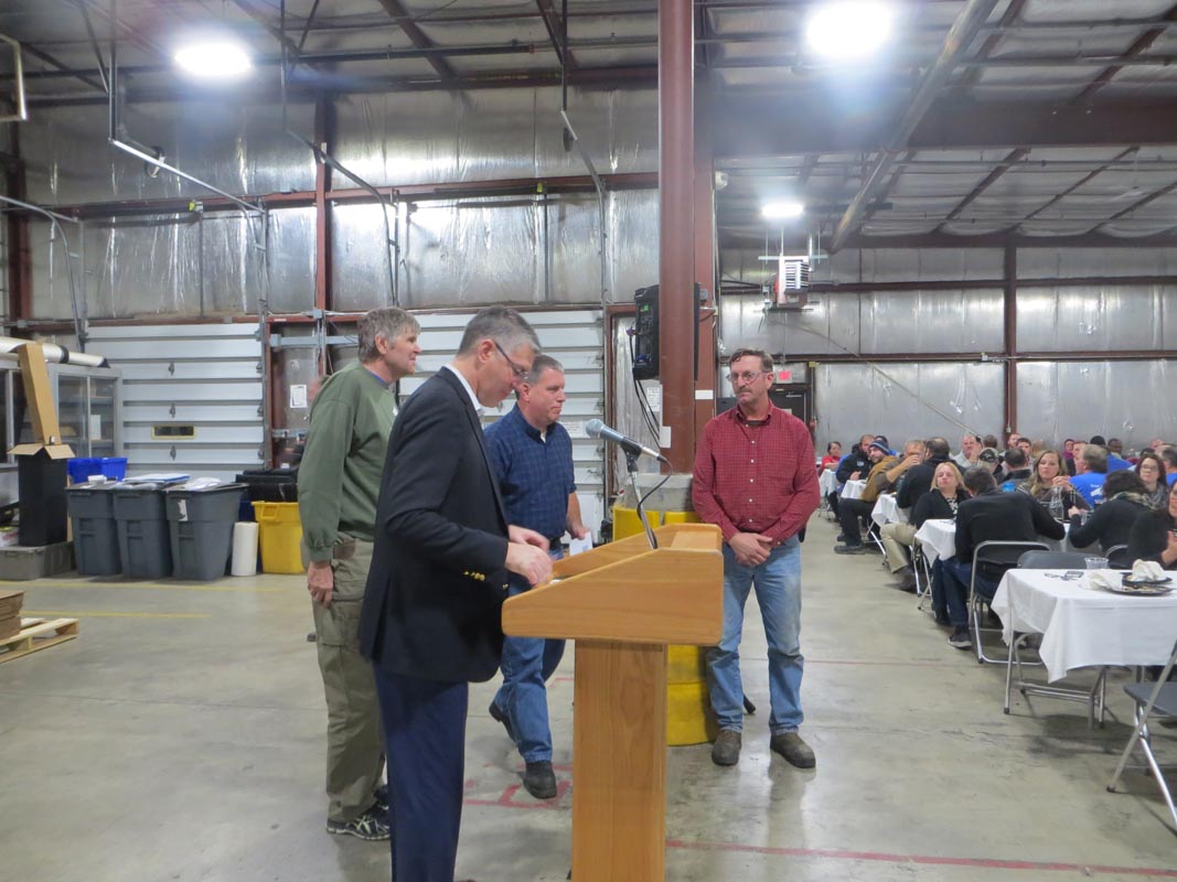 Careers 14 Men gather around a podium as one speaks to an audience seated at tables in an industrial hall