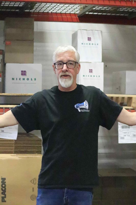 Careers 32 Gray-haired man wearing glasses and a black T-shirt stands with his arms outstretched in a warehouse